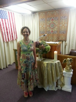 Rev. Sharon Flynn in a print, sleeveless dress, standing in front of an alter with an American flag hanging to the left.